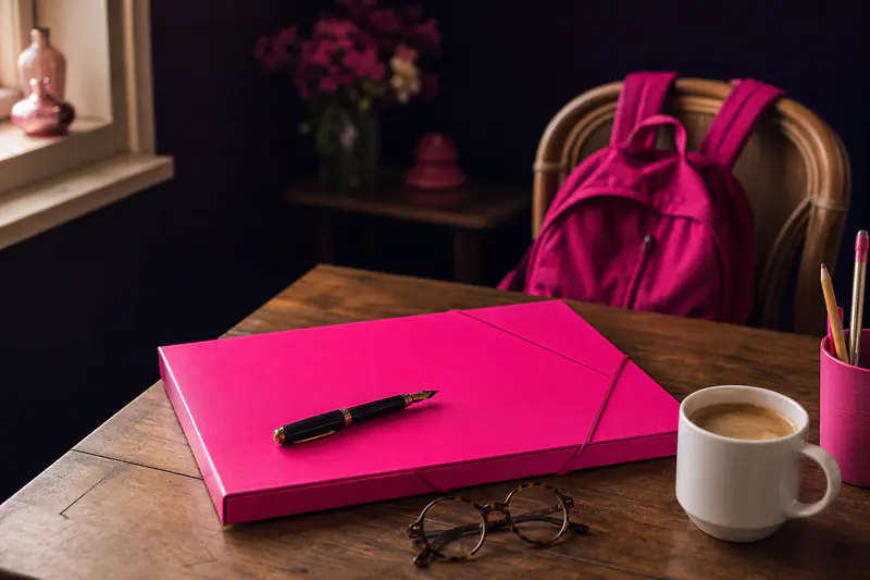 Pochette rose fermée, stylo et lunettes sur une table de cuisine, petit cartable magenta derrière