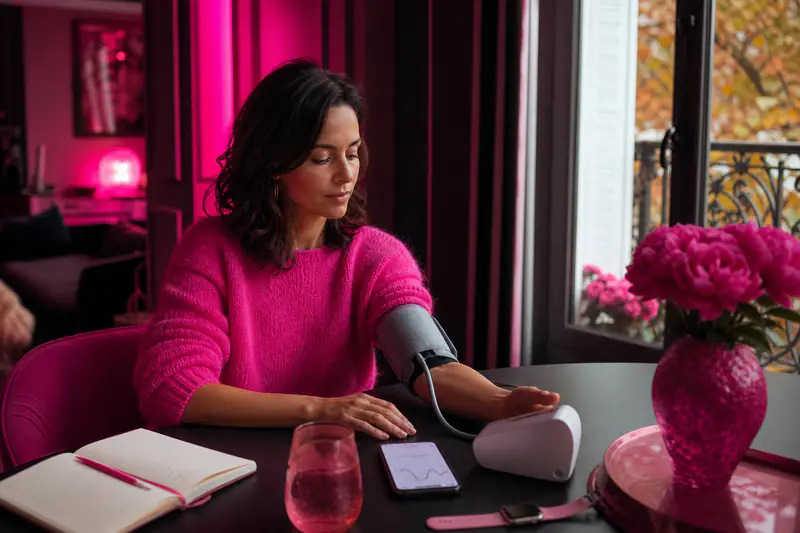 Femme utilisant un tensiomètre connecté avec montre et téléphone sur une table.
