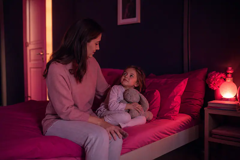Mère assise au bord du lit d’un enfant, veilleuse orangée allumée, porte entrouverte