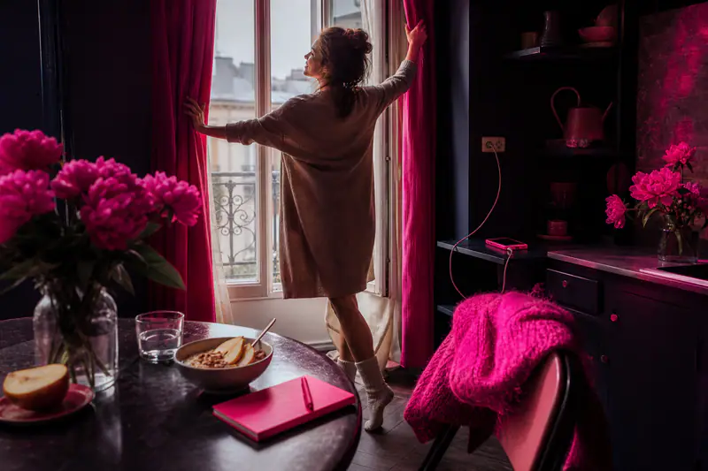 Femme ouvrant ses rideaux avant son petit-déjeuner d’automne dans une cuisine lumineuse.