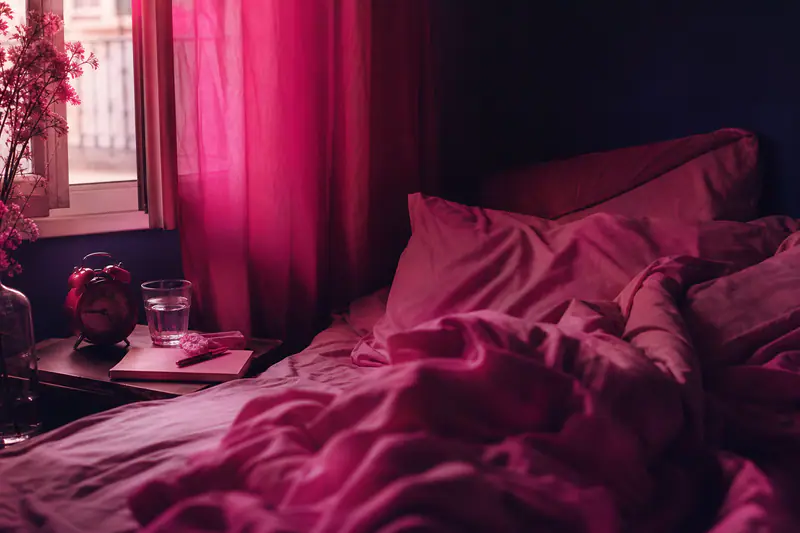 Lit défait au petit matin, réveil, verre d’eau et carnet de sommeil sur la table de nuit
