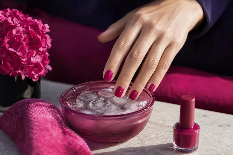 Main aux ongles fraîchement vernis en magenta, au-dessus d’un bol d’eau glacée